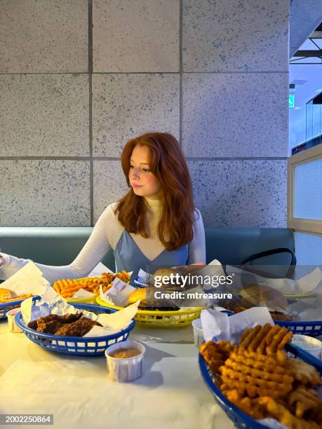 image of attractive, young woman with long, red, wavy hair in diner  restaurant setting, diner table covered with plastic baskets lined with greaseproof paper, smash burgers, potato waffles, chicken tenders, focus on foreground - spaghetti straps stock pictures, royalty-free photos & images