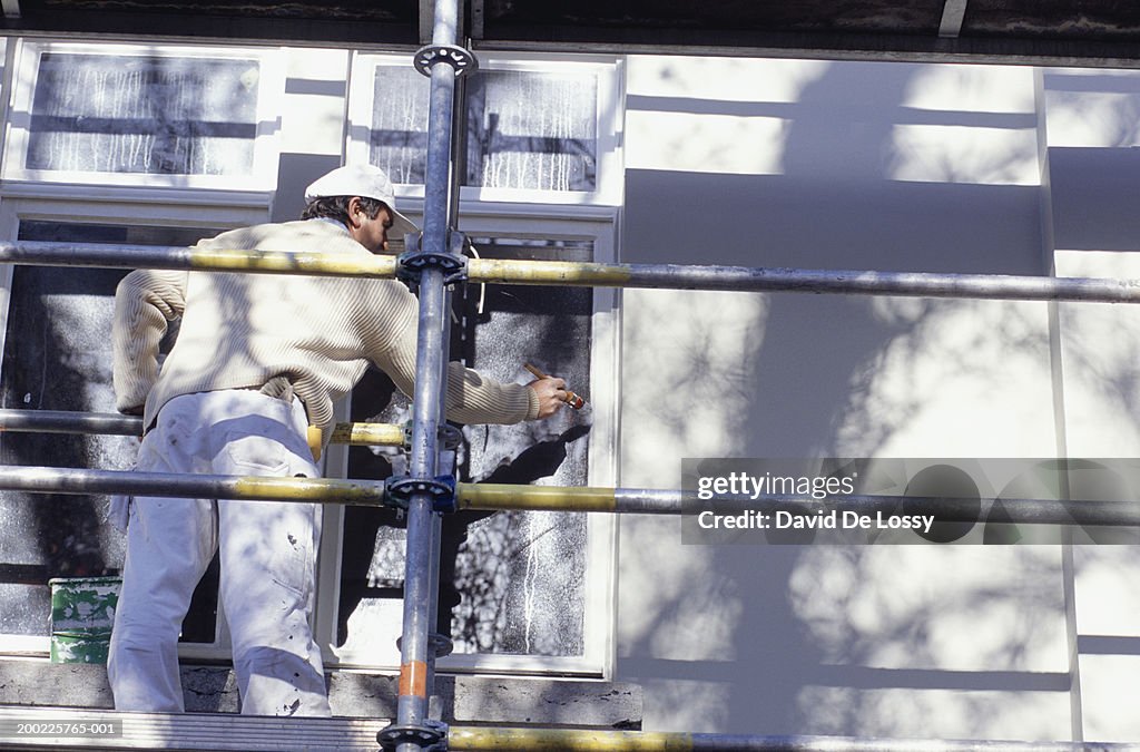 Mature man on scaffolding painting window frames, rear view