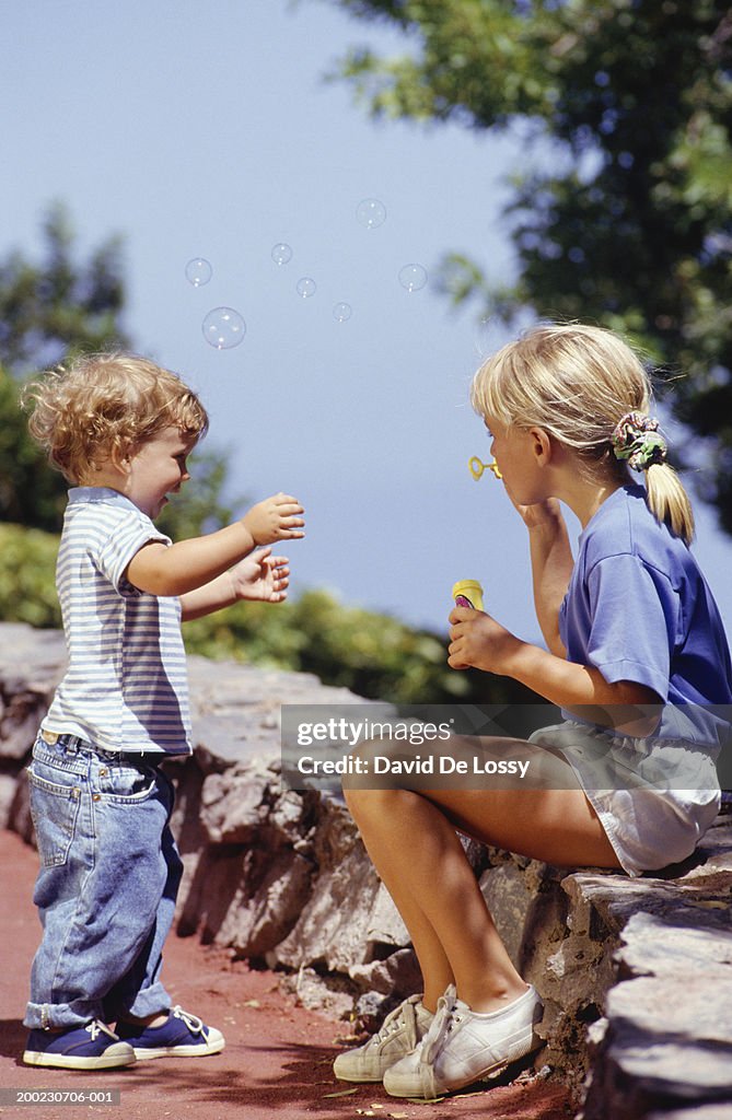 Young girl (8-9) sitting, blowing bubbles with young boy (2-3)