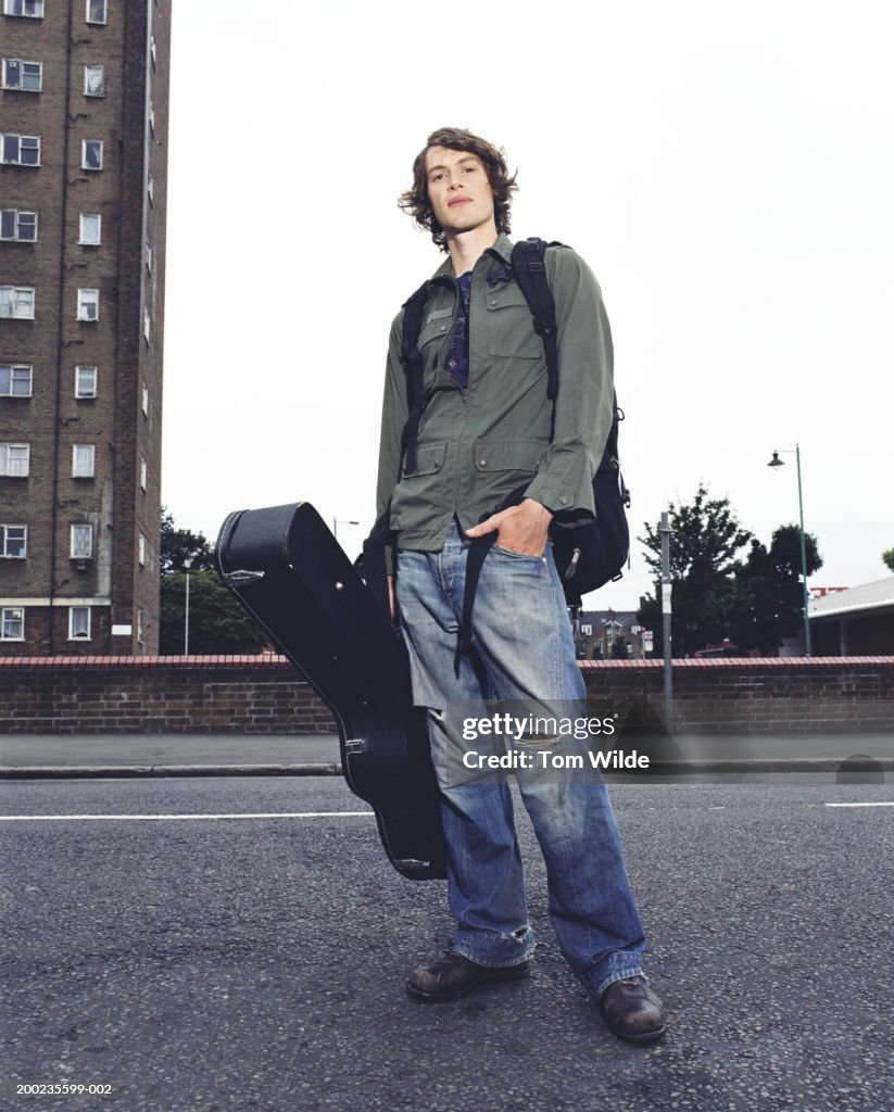 Young man standing in road with rucksack and guitar case, portrait