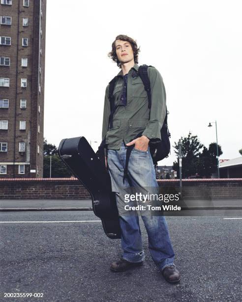 young man standing in road with rucksack and guitar case, portrait - ripped jeans stockfoto's en -beelden