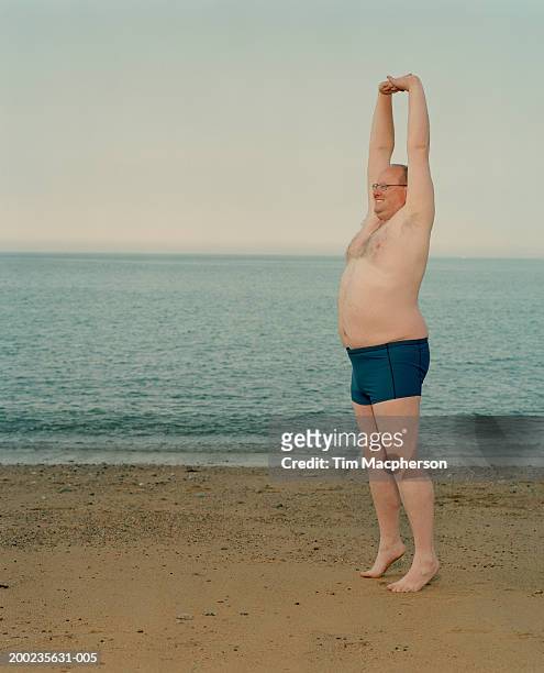 man stretching on beach, standing on tiptoes, arms raised above head - mens swimwear stock pictures, royalty-free photos & images