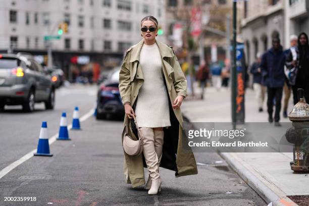 Guest wears sunglasses, golden earrings, a pastel pale green khaki trench long coat , a white ribbed wool mini dress , knee high leather boots , a...