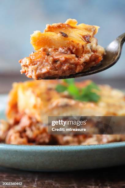 nahaufnahme einer gabel voll hackfleisch und nudeln aus einer portion lasagne auf grün-blauem teller, italienisches essen mit goldener oberfläche, garniert mit flachblättriger petersilie, holzmaserungstischhintergrund, fokus auf vordergrund - golden egg restaurant stock-fotos und bilder