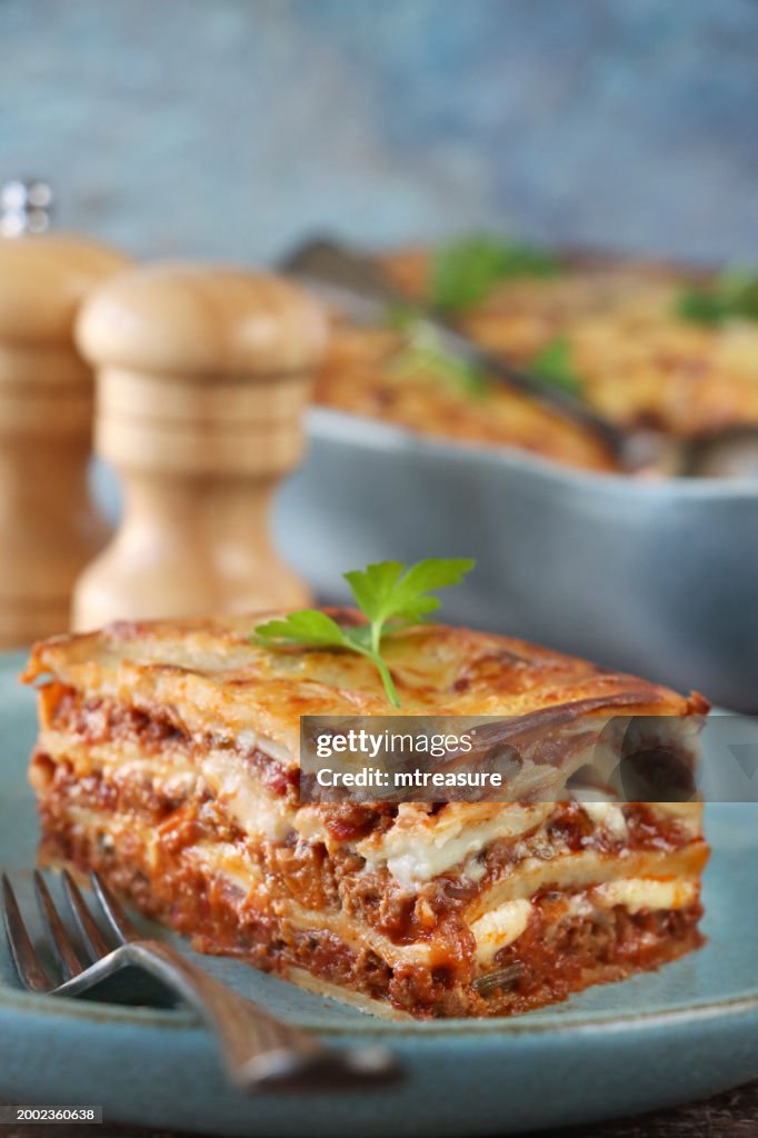 Image of portion of lasagna on green-blue plate with fork, Italian meal of lasagna with missing portion in eggshell blue, ceramic serving dish and spoon, golden surface garnished with flat leaf parsley, salt and pepper shakers, blue background