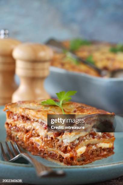 image of portion of lasagna on green-blue plate with fork, italian meal of lasagna with missing portion in eggshell blue, ceramic serving dish and spoon, golden surface garnished with flat leaf parsley, salt and pepper shakers, blue background - selective focus stock pictures, royalty-free photos & images
