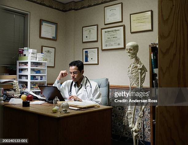 young male doctor using laptop in office, skeleton next to desk - human skeleton stock pictures, royalty-free photos & images