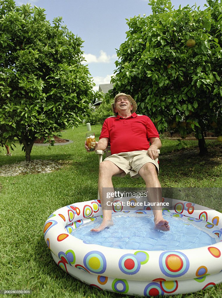 Senior man dangling feet in kiddie pool, holding drink