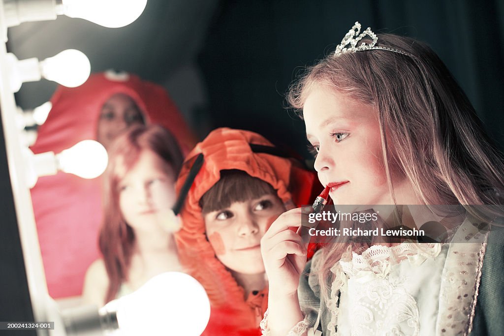Two girls (5-8), one watching friend applying lipstick in stage mirror