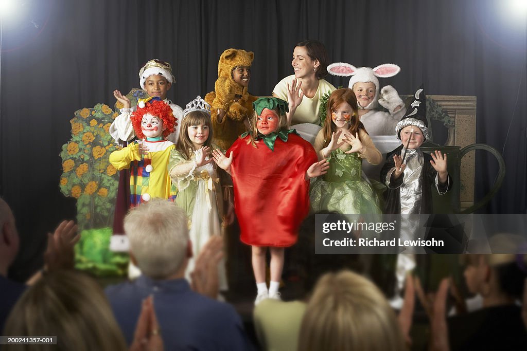 Crianças (4-9) vestindo figurinos e professor acenando no palco
