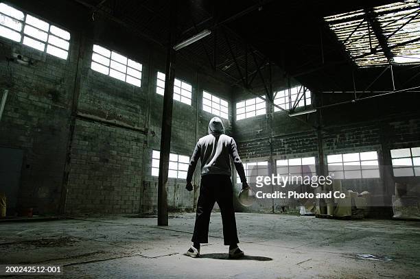 man wearing hooded jacket, holding football in warehouse, rear view - uomo incappucciato foto e immagini stock