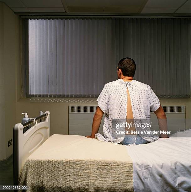 young man wearing hospital gown, sitting on bed, rear view - man in hospital stock pictures, royalty-free photos & images