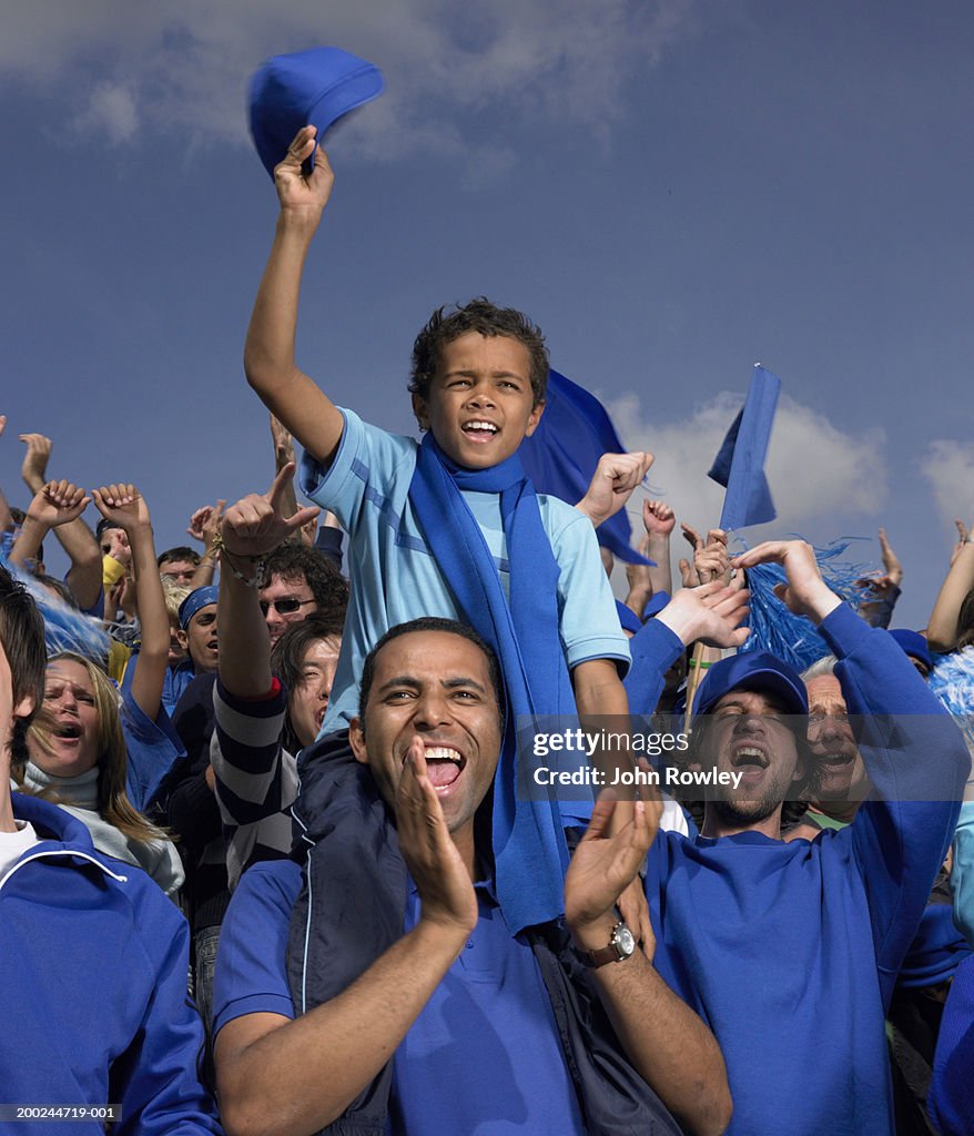Stadium crowd cheering, boy (5-7) on father's shoulders, waving cap