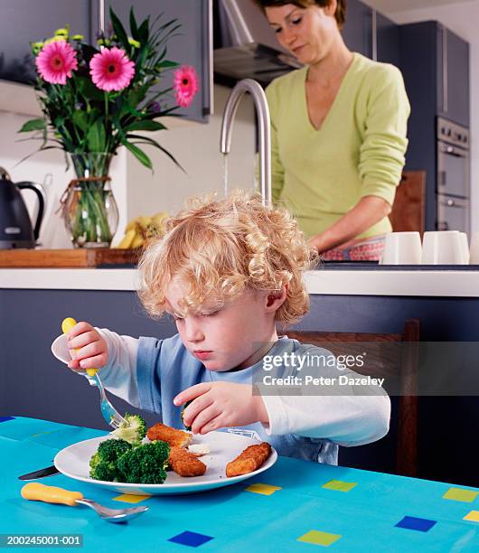 boy (2-4) eating fish fingers and broccoli, mother at kitchen sink - fish fingers stockfoto's en -beelden