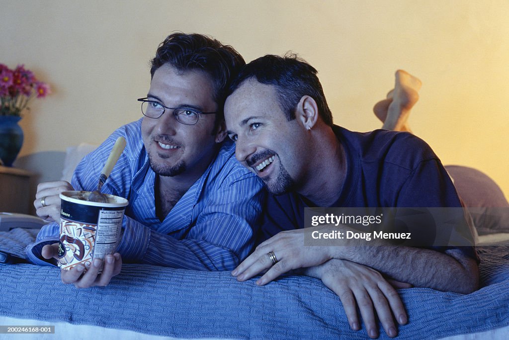 Gay couple lying on bed watching television, one eating ice cream
