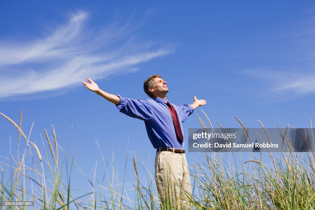 Mature businessman standing in field, arms outstretched, eyes closed