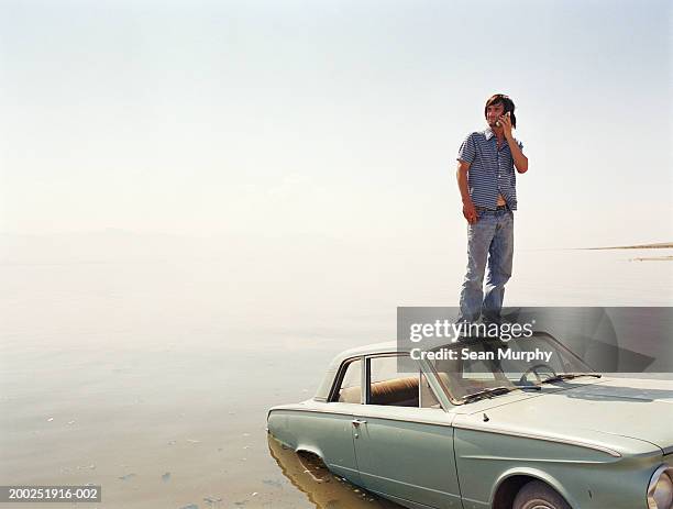 young man standing on roof of car in water, using mobile phone - salton sea stock pictures, royalty-free photos & images