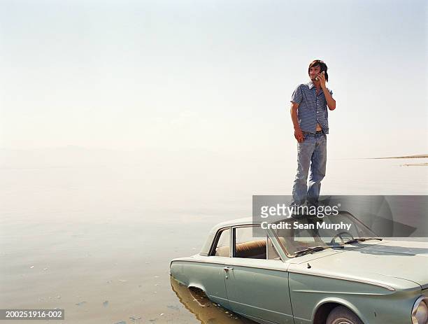 young man standing on roof of car in water, using mobile phone - sunken car bildbanksfoton och bilder