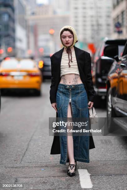 Guest wears a yellow knitted wool balaclava, a black coat , red lipstick , make-up , a gathered crop top, tattoos, a blue denim slit skirt , pointed...