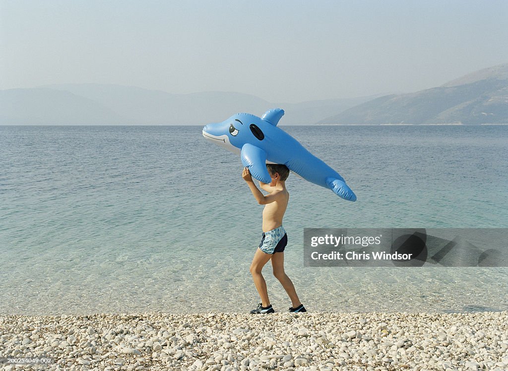 Boy (8-10) on pebbled beach with inflatable dolphin on head, side view