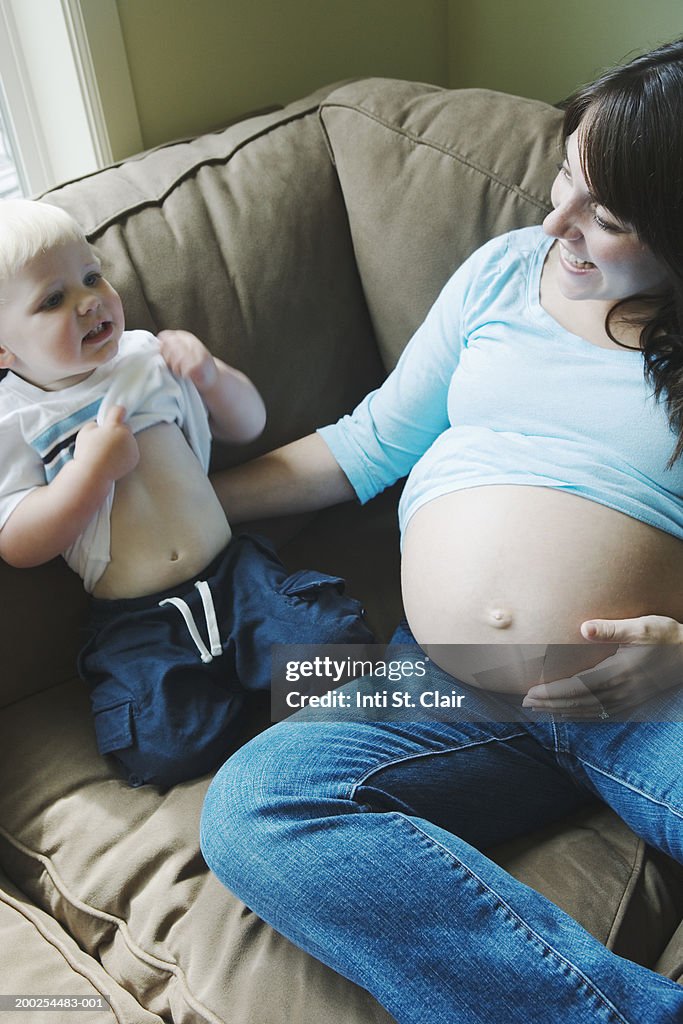 Pregnant woman and son (2-4) sitting on sofa, boy lifting shirt