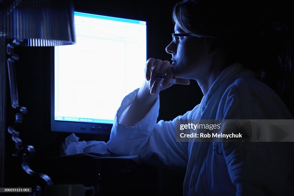 Woman in robe using computer in dark room, resting chin on hand