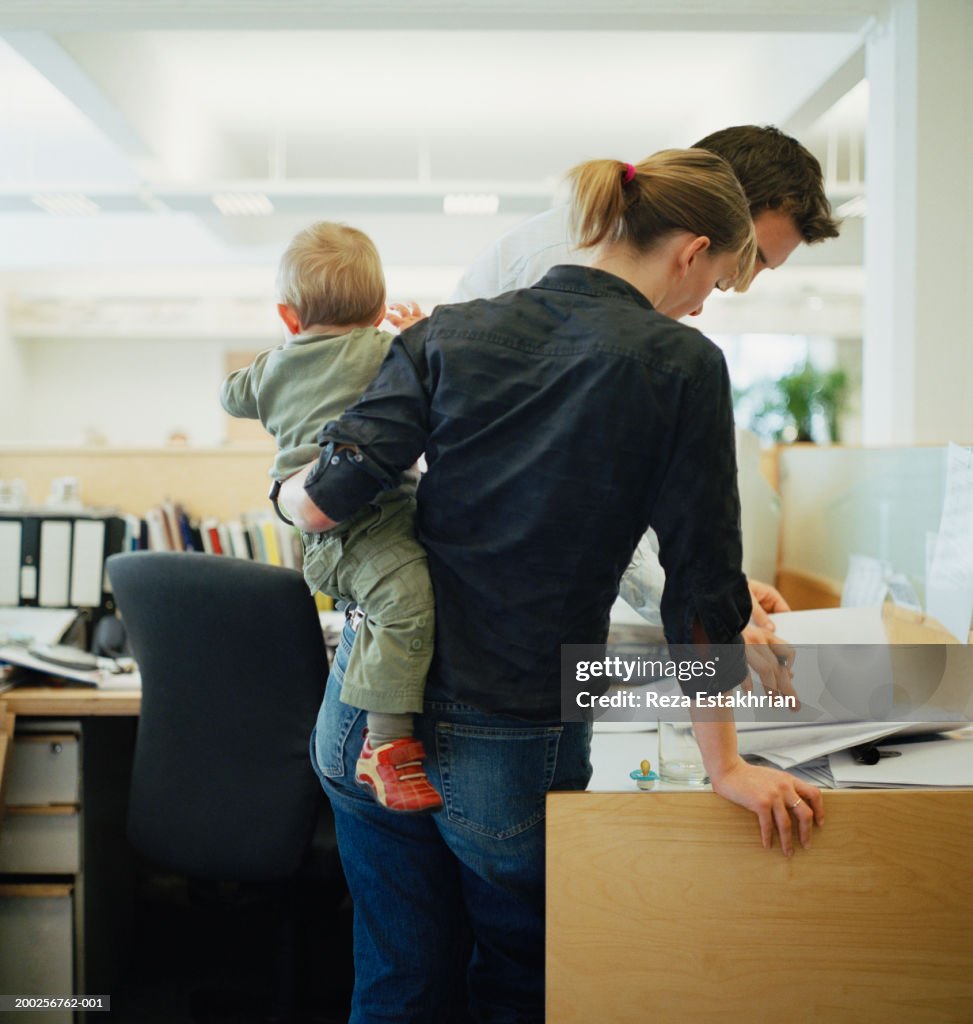 Young woman holding toddler boy, talking to young man in office