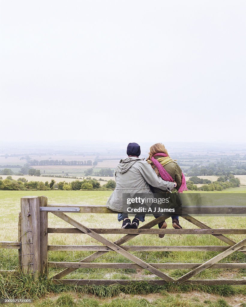 Young couple embracing, sitting on fence by field, rear view