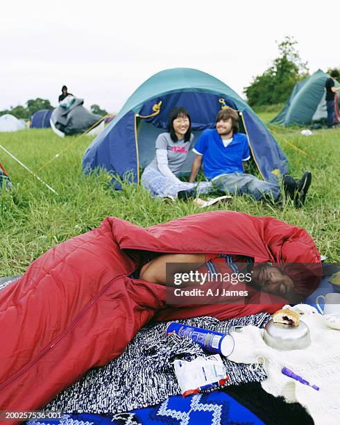 young man lying in sleeping bag, couple by tent in background - festival camping stock pictures, royalty-free photos & images