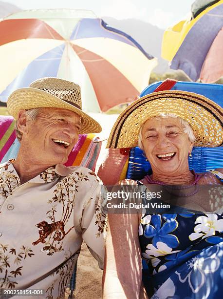 senior couple sitting on beach under umbrella, laughing - beach hat stock pictures, royalty-free photos & images