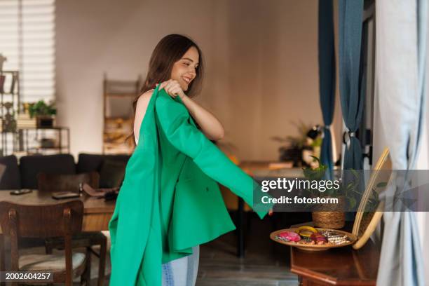 mulher nova olhando no espelho e colocando um blazer verde - blazer - fotografias e filmes do acervo
