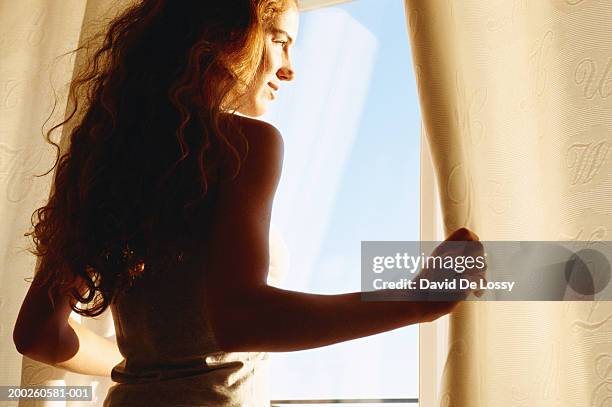 young woman smiling, looking out window - window curtains stock pictures, royalty-free photos & images