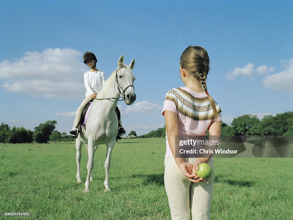 Girl (6-8) holding apple behind back, facing sister (10-12) on horse