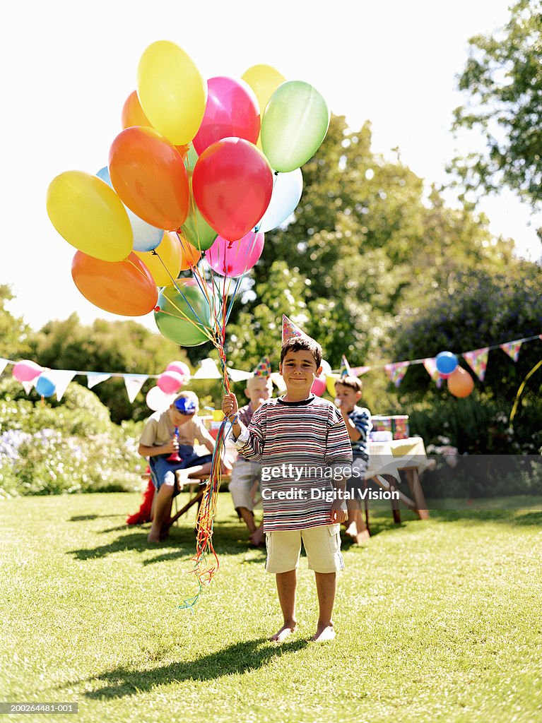 Boy (5-7) holding bunch of balloons outdoors, friends in background