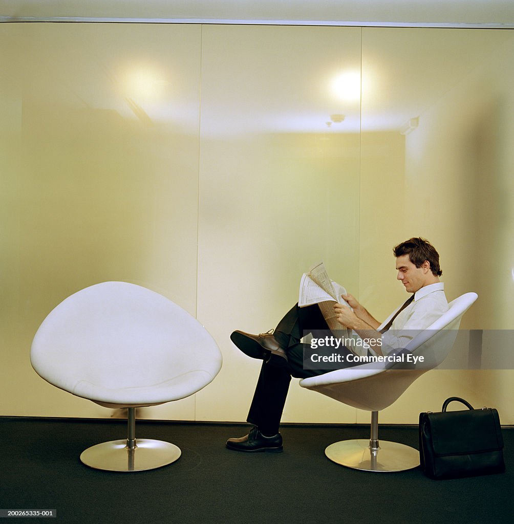 Businessman sitting on chair, reading newspaper, side view