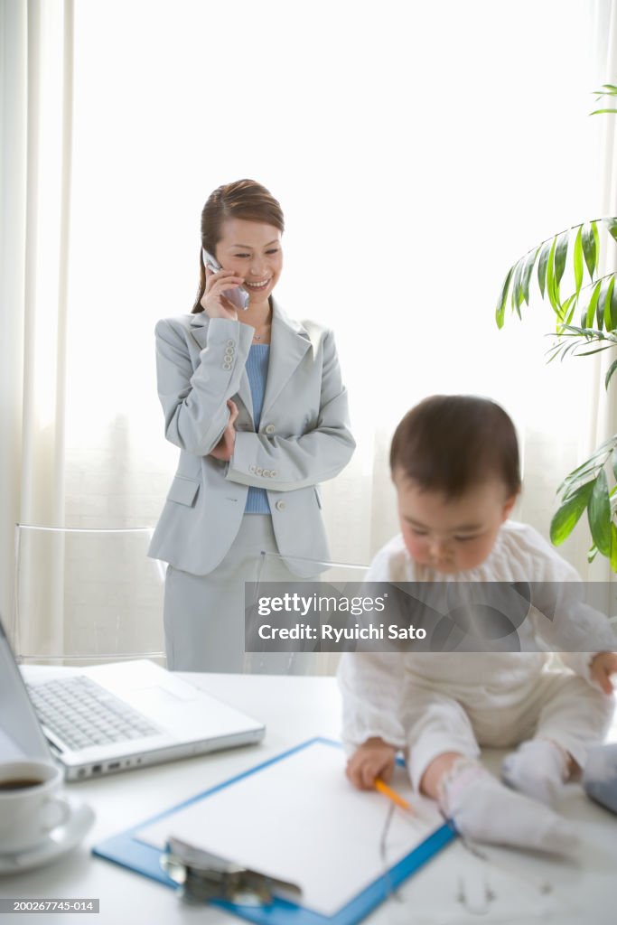 Mother using mobile phone, daughter (6-9 months) sitting on desk