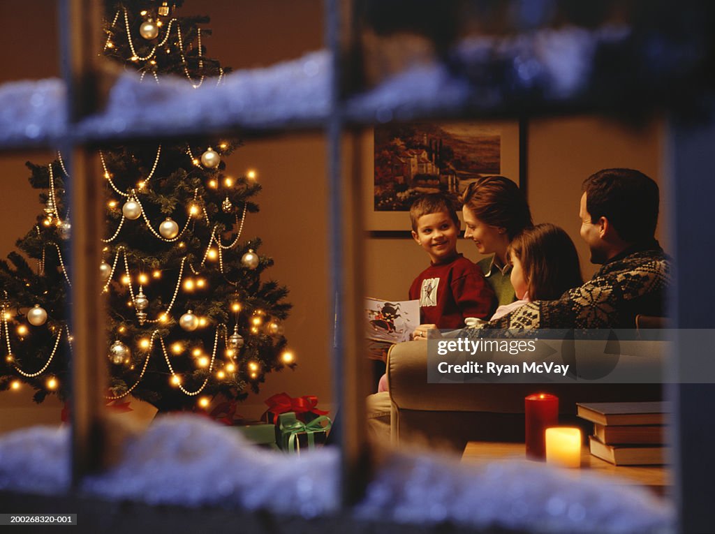 Family reading together on sofa at Christmas time, viewed through window