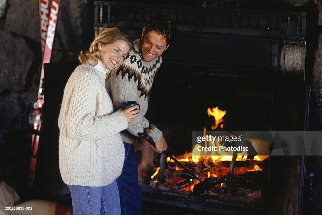 Couple in ski lodge standing  by fireplace, man stoking fire