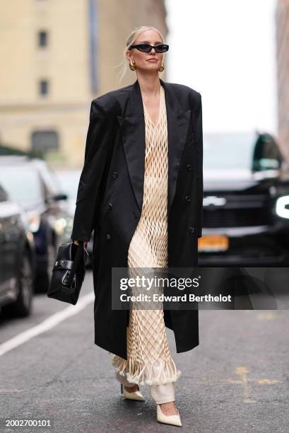 Leonie Hanne wears sunglasses, golden earrings, a black long blazer jacket / coat, a leather bag, a pastel pale yellow mesh fishnet long dress with...