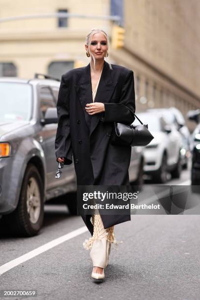 Leonie Hanne wears golden earrings, a black long blazer jacket / coat, a leather bag, a pastel pale yellow mesh fishnet long dress with v-neck /...