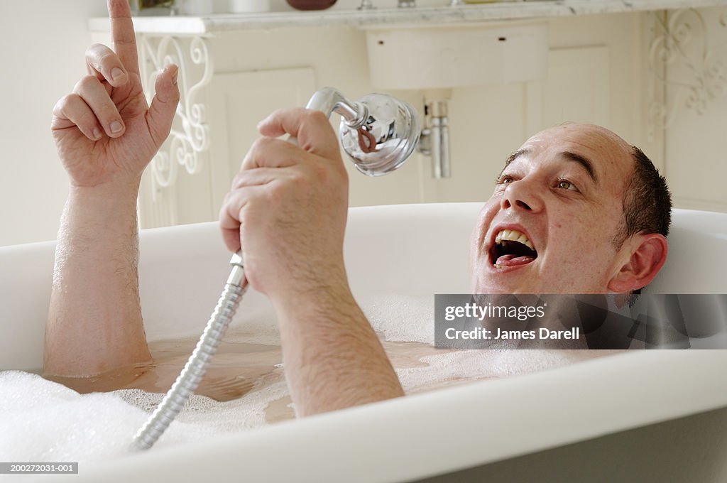 Man lying in bath singing into shower head, close-up