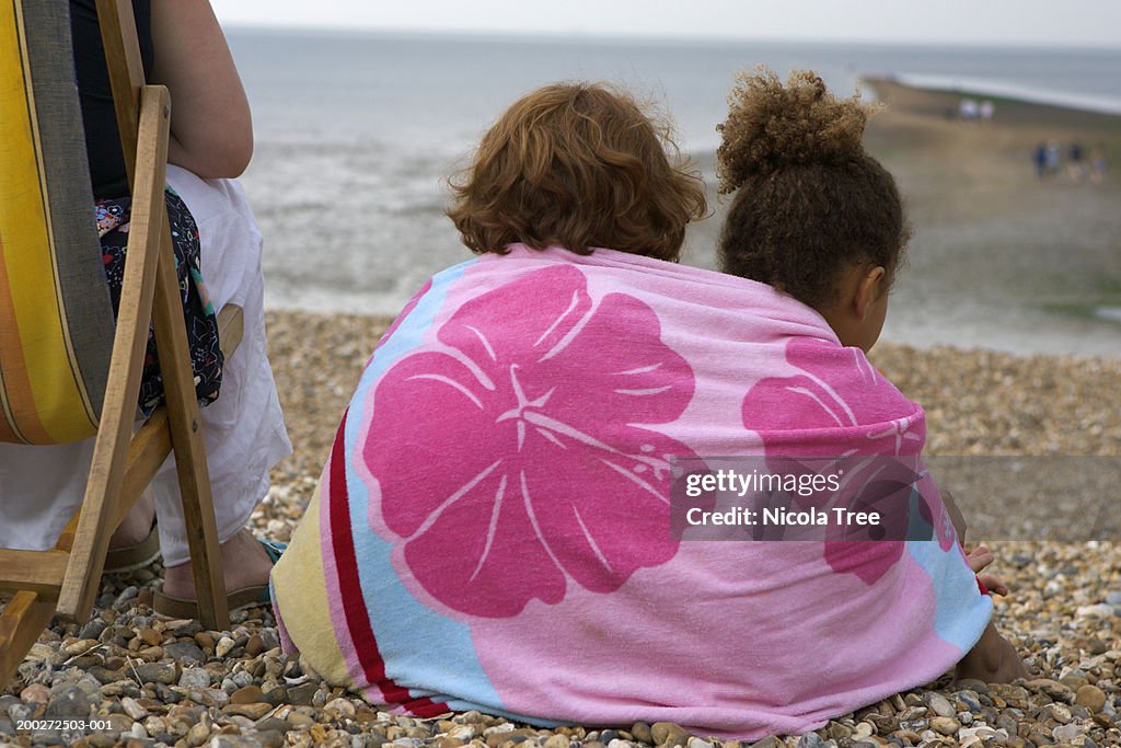 Two girls (6-8) sitting on pebbled beach beneath towel, rear view