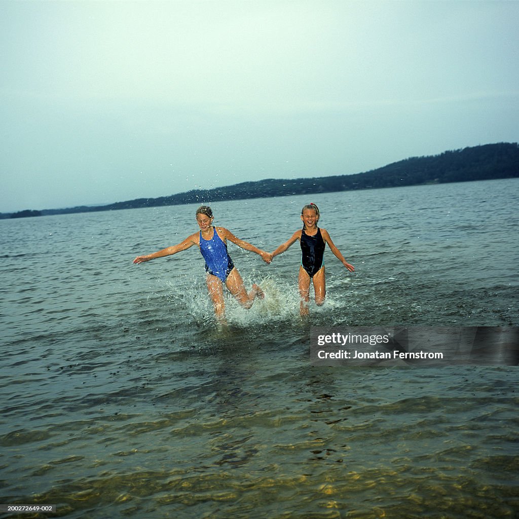 Two sisters (9-11) holding hands splashing in lake, smiling