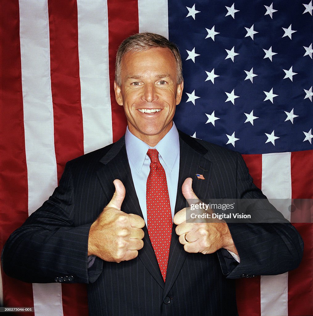 Businessman giving thumbs-up sign in front of American flag, smiling