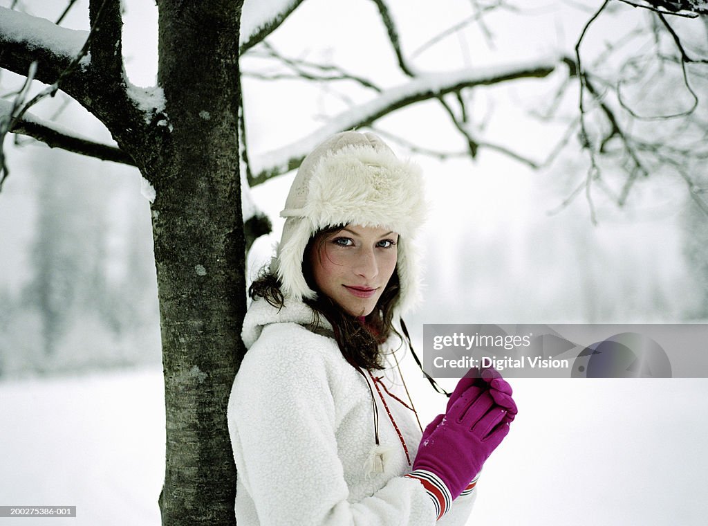 Young woman standing in snow, leaning against tree, portrait