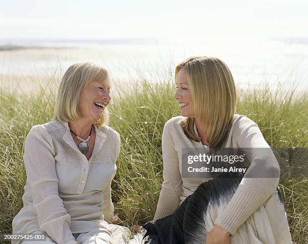 mother and mature daughter sitting on grass by sea, smiling - abbracciare le ginocchia foto e immagini stock