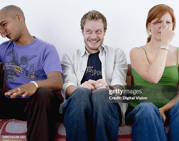 young man, smiling, sitting between man and woman holding noses - farting fotografías e imágenes de stock