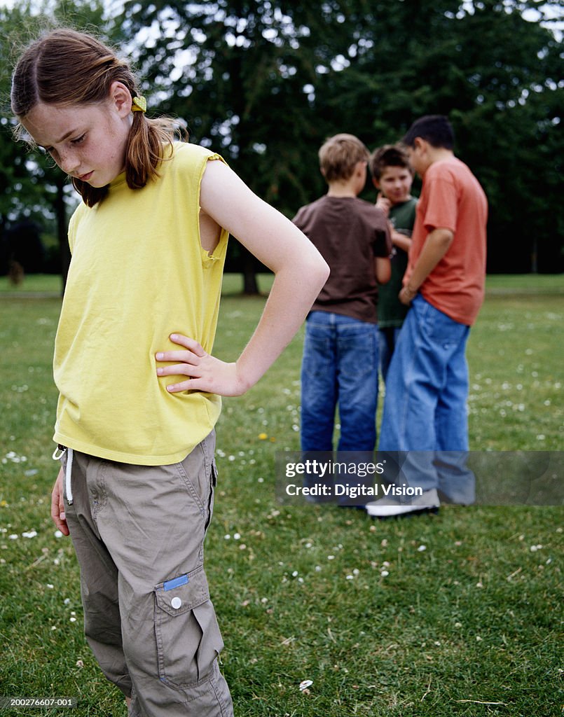 Girl (9-11) in park hand on hip, looking down, boys (9-12) in background