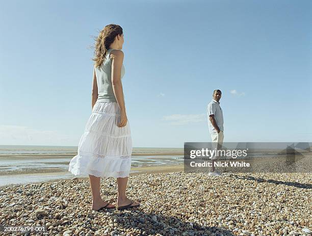 couple on pebble beach, woman looking at man with hands in pockets - white skirt foto e immagini stock