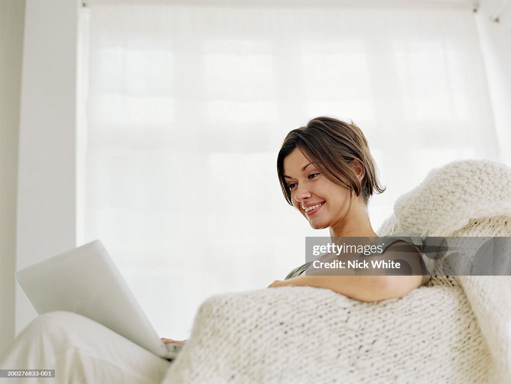 Woman relaxing in armchair looking at laptop computer, smiling